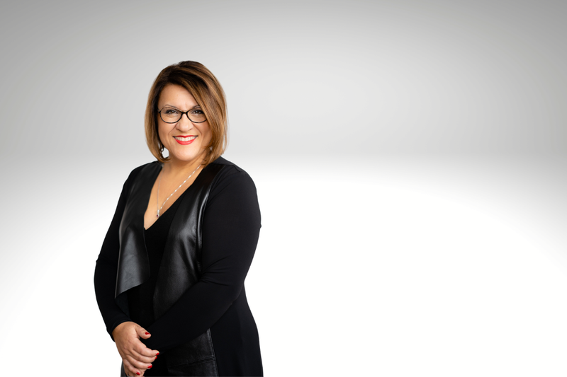 Portrait professionnel d’une femme souriante portant des lunettes, vêtue de noir, posant devant un fond dégradé clair.